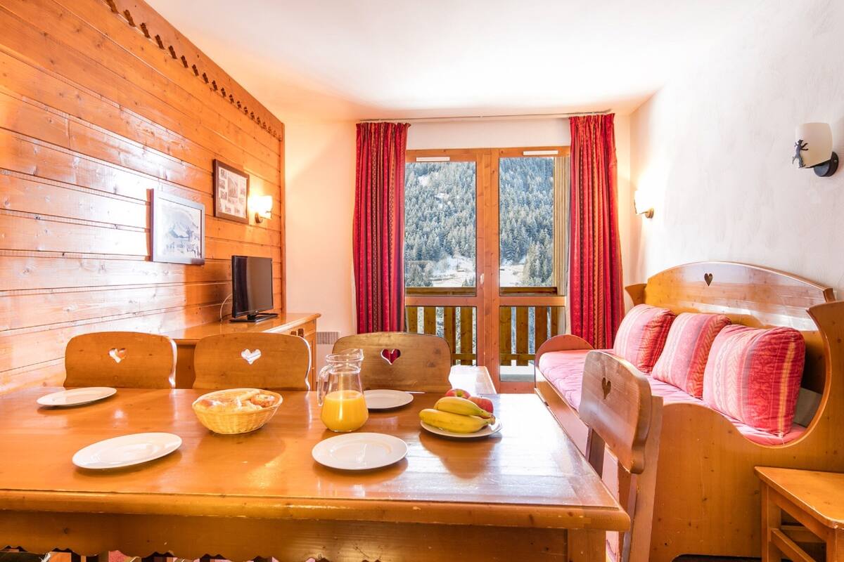 A wooden cabin with a dining table set for breakfast, a bed with pink bedding, and a window with a snowy view.