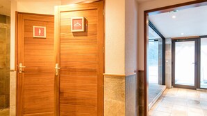 Un conjunto de puertas dobles de madera con un escudo de armas y un símbolo de perchero, que dan paso a un corredor con baldosas y una puerta de vidrio.