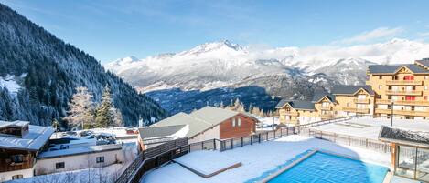 Zona de piscina cubierta de nieve con montañas al fondo, edificios de madera y cielo despejado.