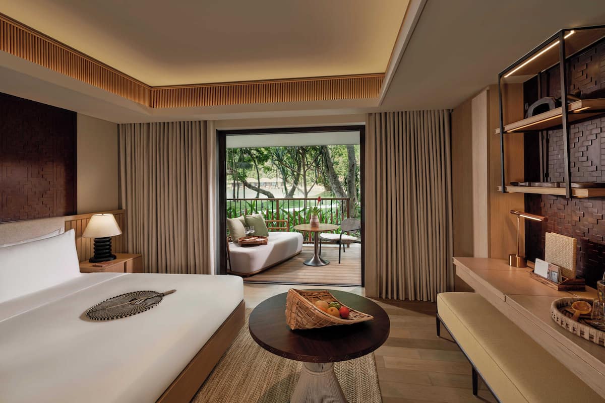 Habitación de hotel moderna con una cama grande, una pequeña mesa redonda con fruta, un sofá y un balcón con vistas a la vegetación.