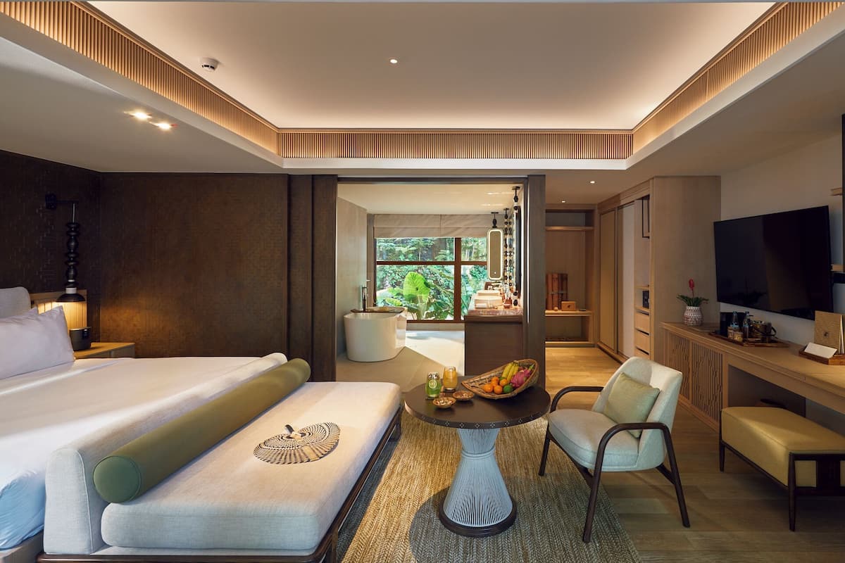 Una habitación de hotel moderna con una cama grande, una zona de estar con una mesa, un televisor y vistas al exterior.