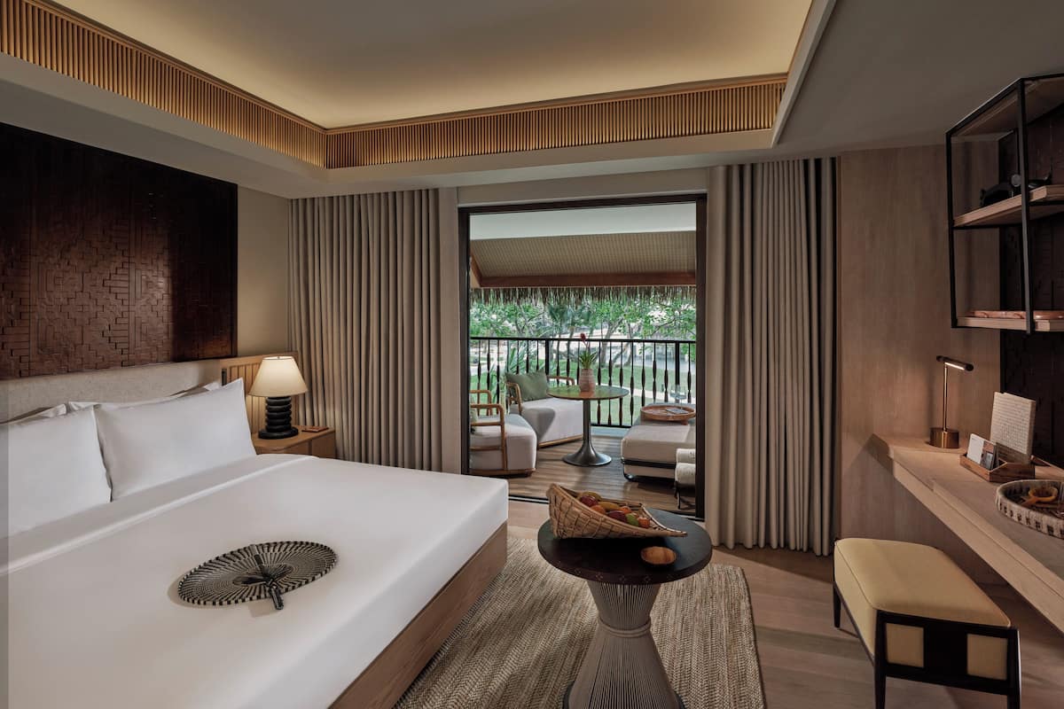 Una habitación de hotel moderna con una cama grande, una mesita con una cesta de frutas, un área de descanso con una mesa y sillas, y un balcón con vistas a zonas verdes.