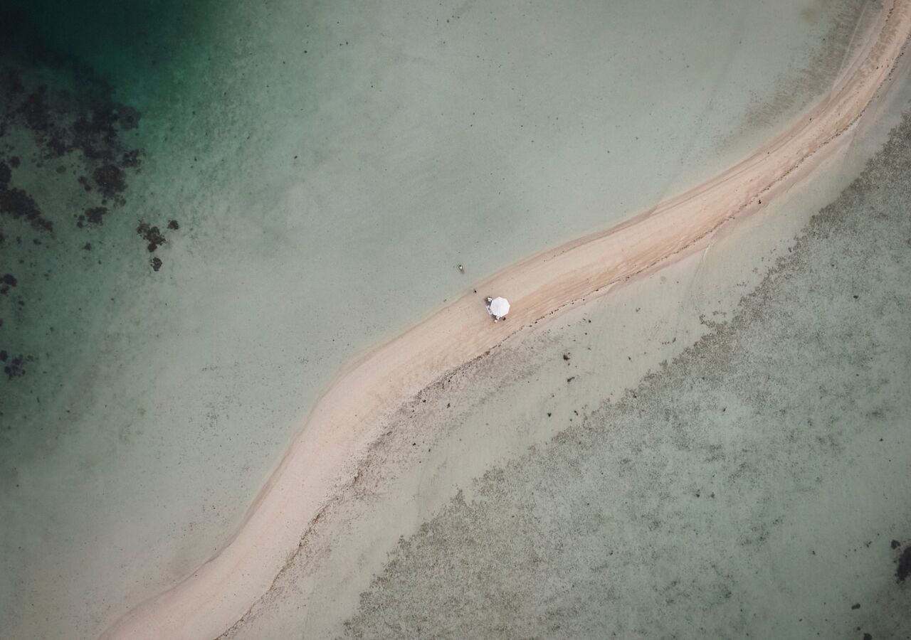 Eine Luftaufnahme eines Strandes mit einem schmalen Sandweg, der zu einem weißen Gegenstand führt, möglicherweise einem Boot oder einem Bojen. Ruhiges Wasser und ein sandiger Uferbereich sind ebenfalls zu sehen.