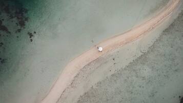 Eine Luftaufnahme eines Strandes mit einem schmalen Sandweg, der zu einem weißen Gegenstand führt, möglicherweise einem Boot oder einem Bojen. Ruhiges Wasser und ein sandiger Uferbereich sind ebenfalls zu sehen.