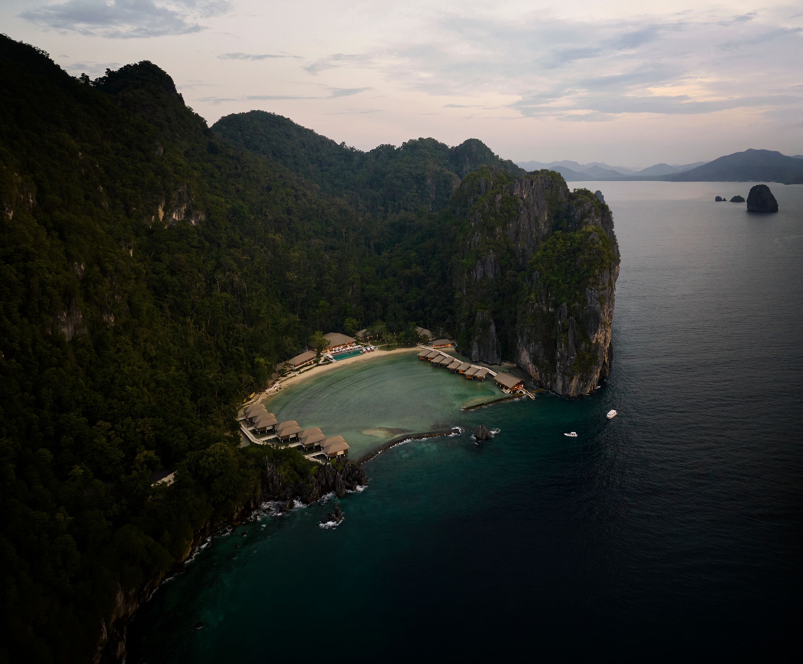 An aerial view of a coastal resort with a beach, thatched-roof structures, and a clear view of the surrounding cliffs and water.