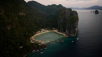 An aerial view of a coastal resort with a beach, thatched-roof structures, and a clear view of the surrounding cliffs and water.