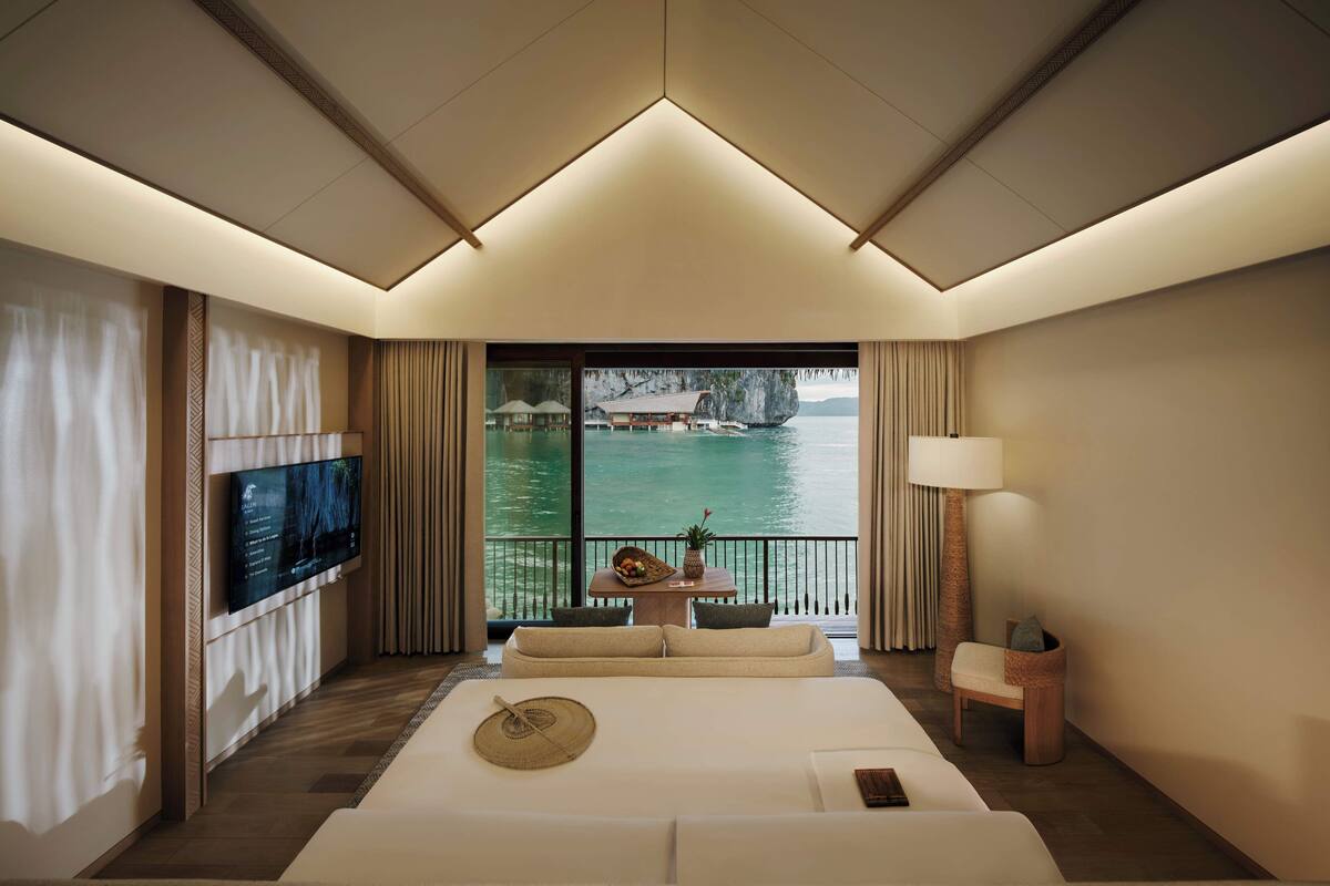 Habitación de hotel con una cama grande, televisión, balcón con vista al mar y a la montaña, y una mesita con lámpara.