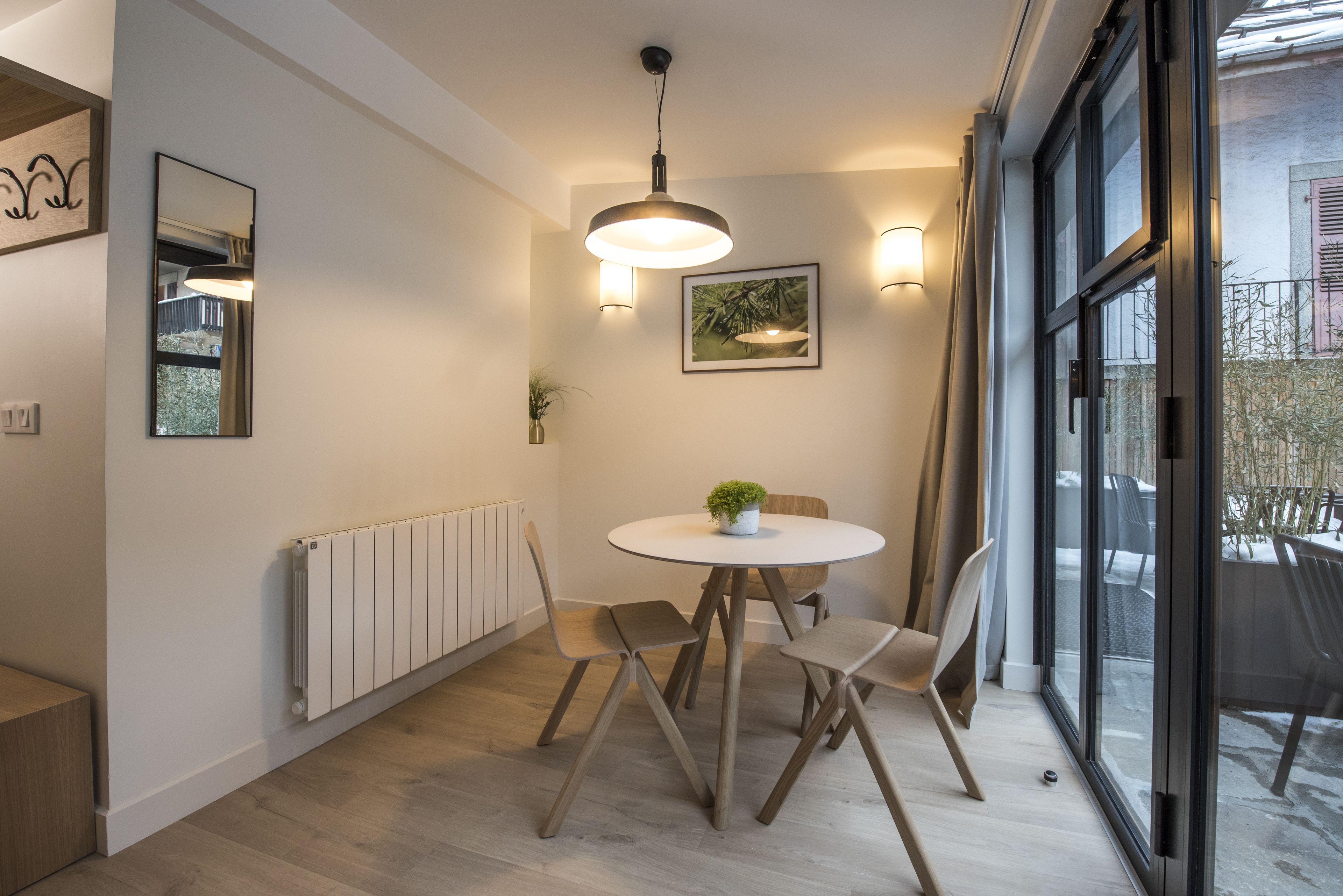 A modern dining area with a round table, two chairs, a pendant light, a framed picture, and a potted plant.