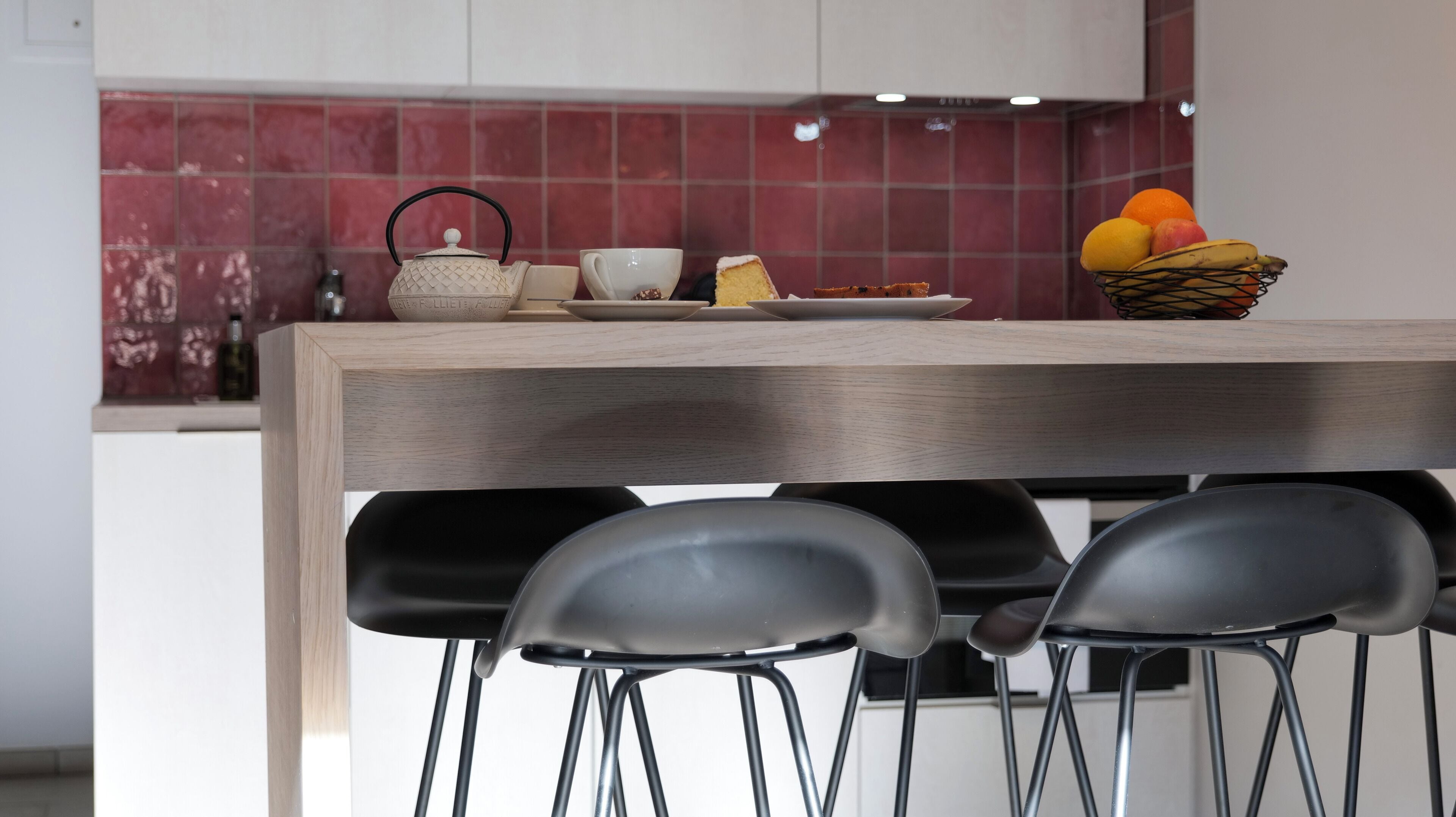 A modern kitchen with a wooden countertop, a kettle, a cup, a plate with food, and a fruit basket. There are three black chairs in front of the counter.