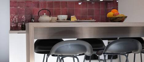 A modern kitchen with a wooden countertop, a kettle, a cup, a plate with food, and a fruit basket. There are three black chairs in front of the counter.