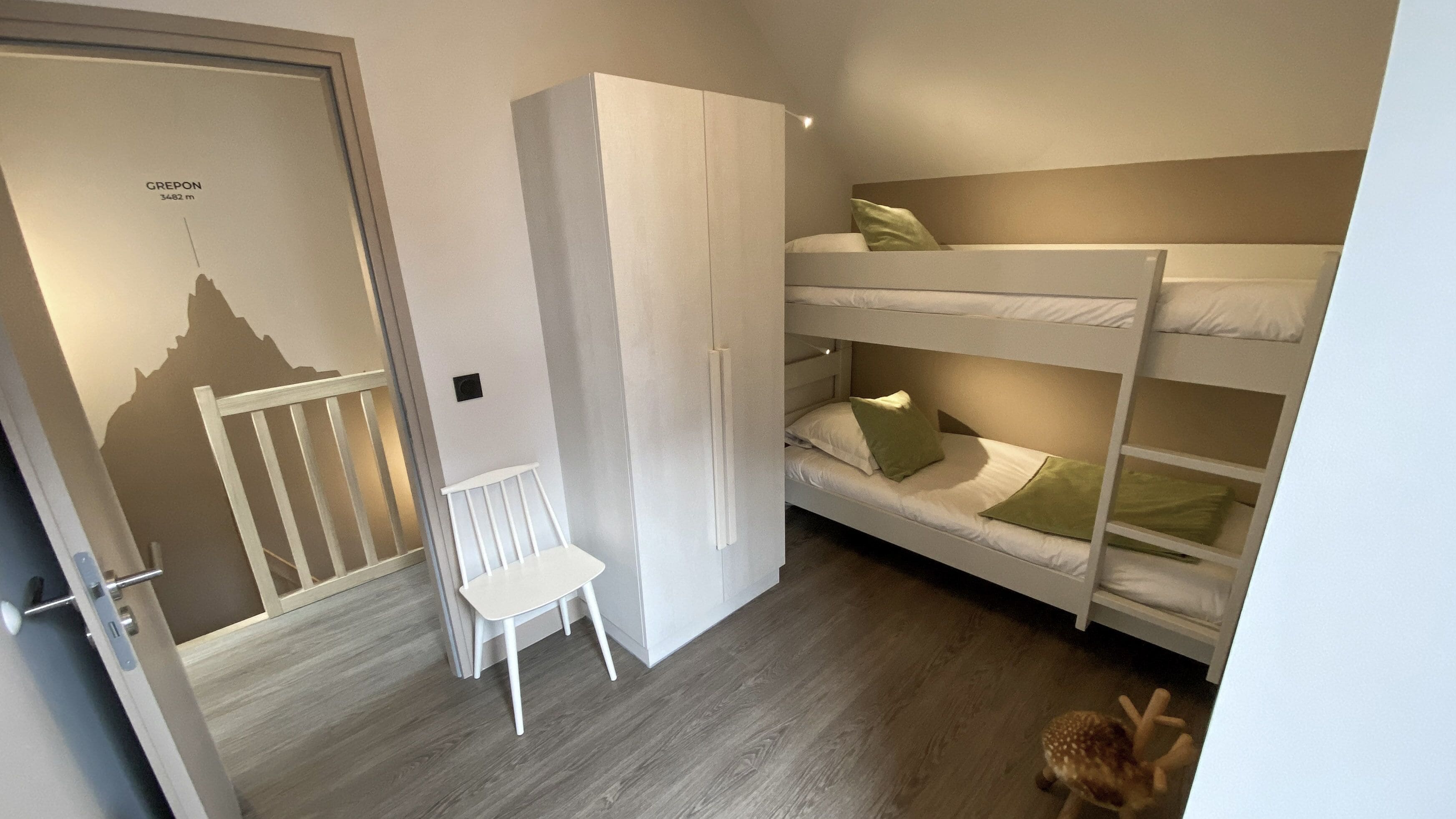 Un dormitorio con una cama litera, un armario blanco, una pequeña silla blanca y una puerta con el silueta de una montaña.
