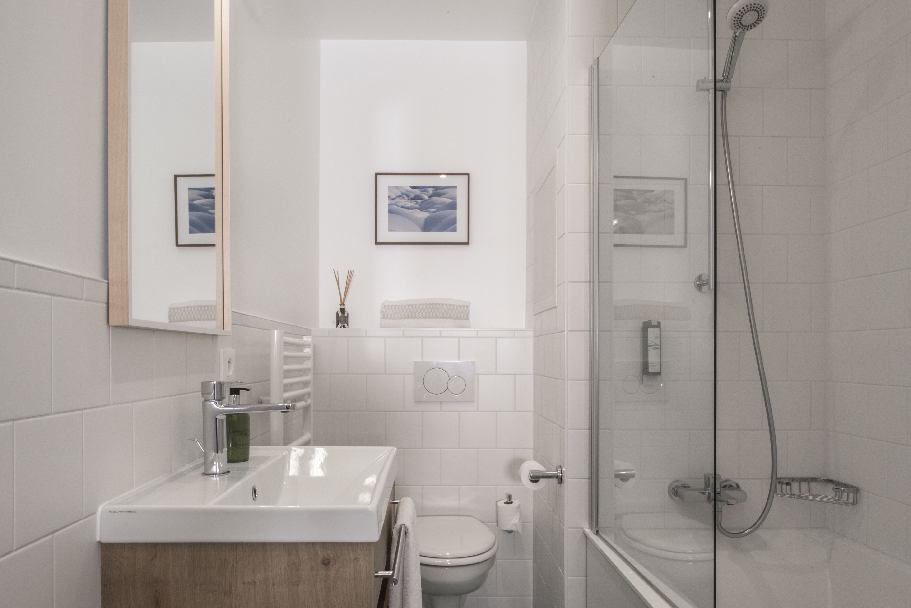 A modern bathroom with a white sink, toilet, and shower. There are framed pictures on the wall and a mirror above the sink.