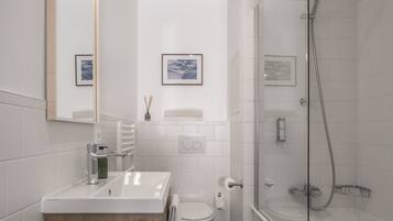 A modern bathroom with a white sink, toilet, and shower. There are framed pictures on the wall and a mirror above the sink.