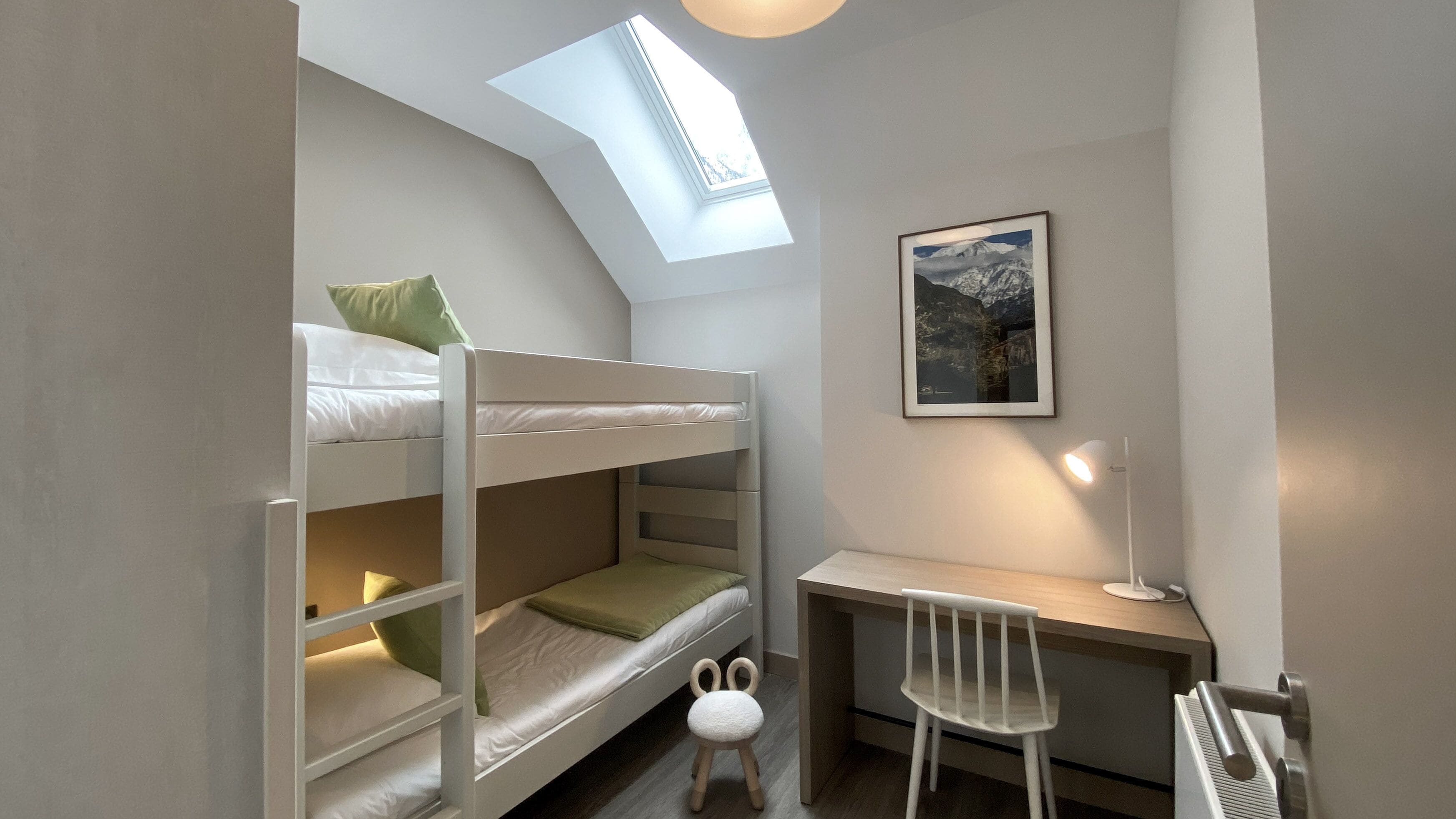 A dormitory room with bunk beds, a desk, a chair, a radiator, and a skylight.