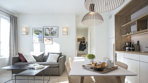 A modern living room with a sofa, coffee table, and dining table. There is a kitchen area with a coffee machine and a plant. - Appart'Hôtel le Génépy (Chamonix-Mont-Blanc)