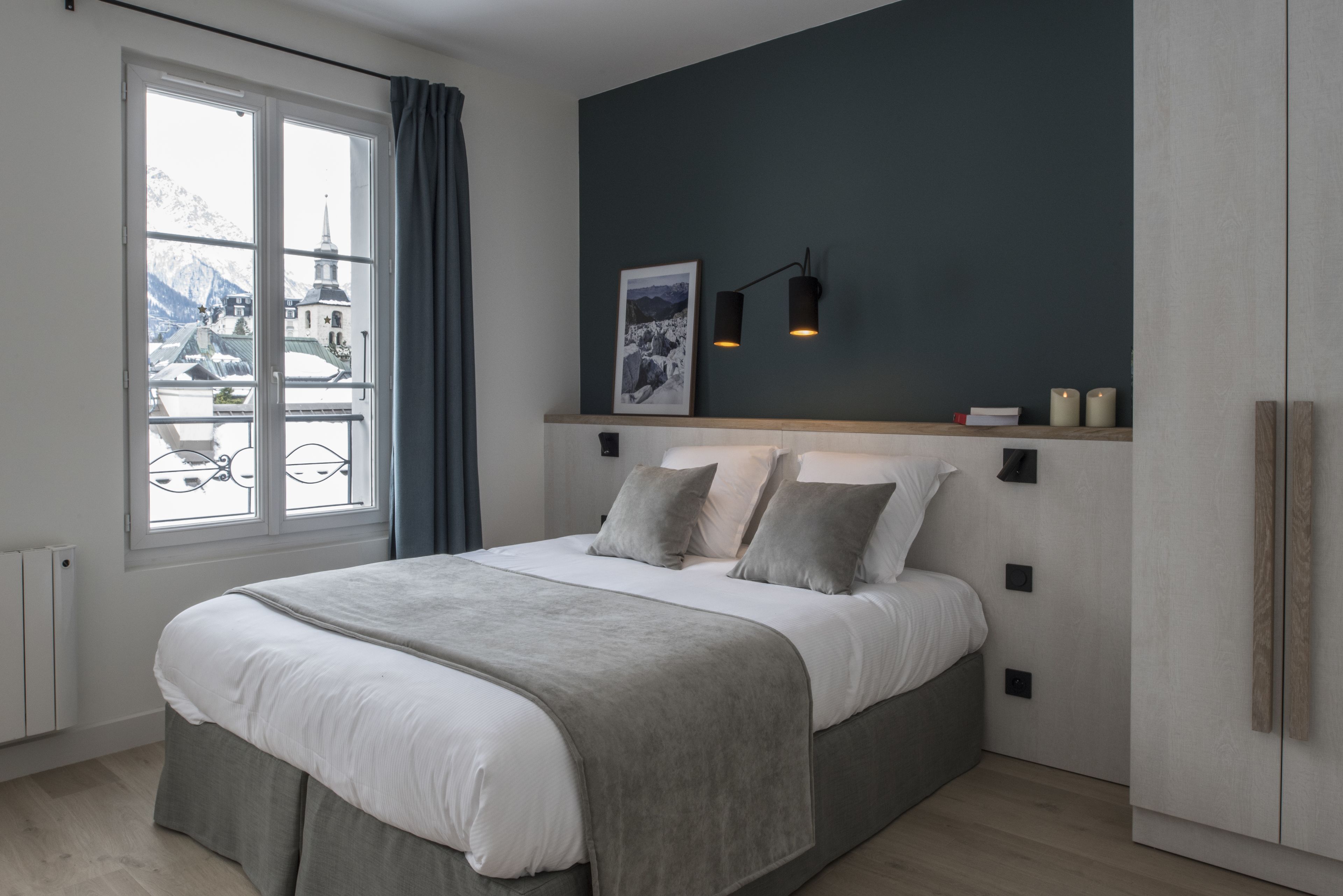 Ein modernes Schlafzimmer mit einem großen Bett, einem Fenster mit Blick auf Berge, einer dunklen Wand und einem Regal mit Büchern und Kerzen.