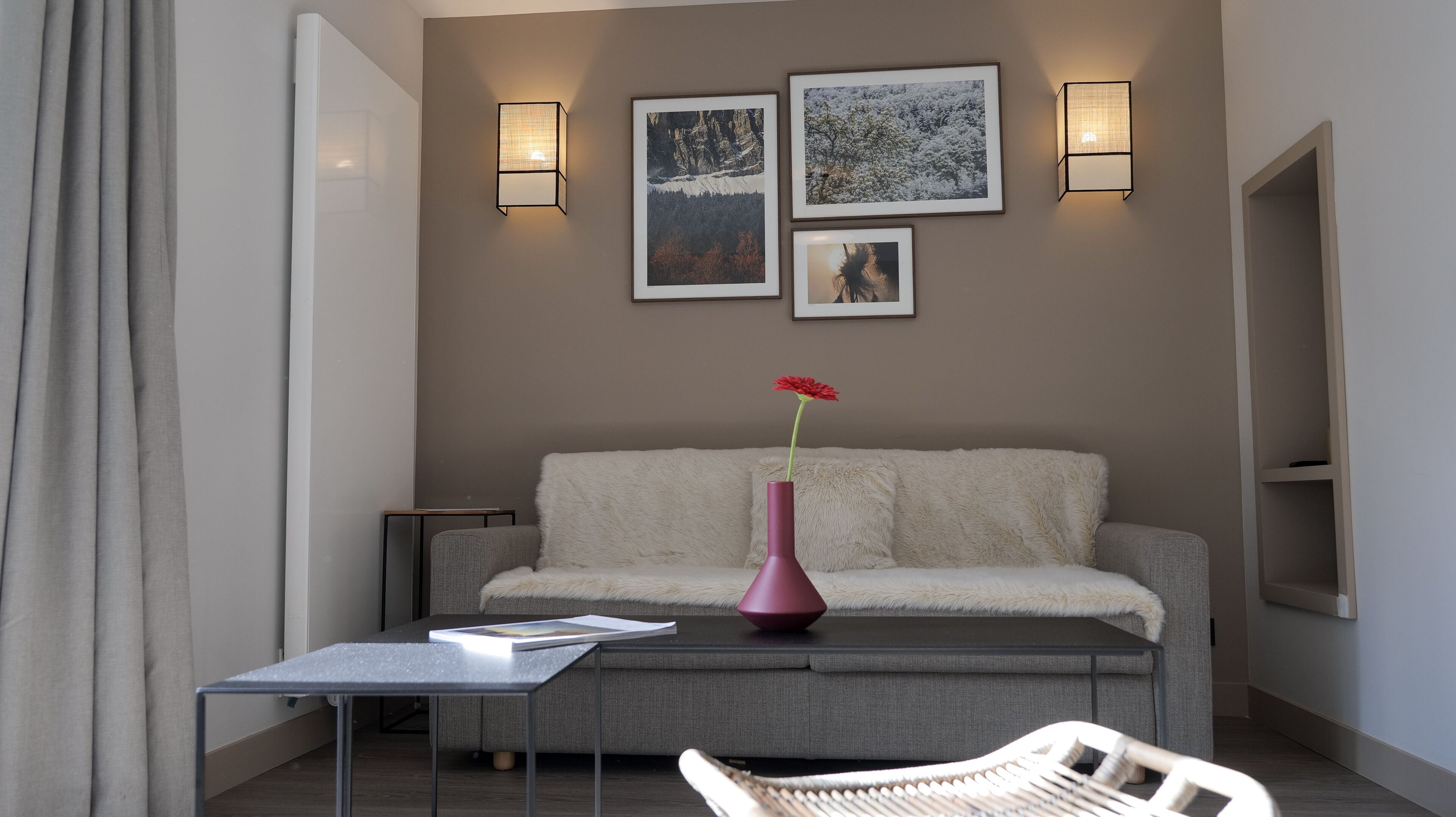 Een moderne woonkamer met een bank, een salontafel en een vaas met een bloem. Er zijn wandlampen en ingelijste foto's aan de muur.