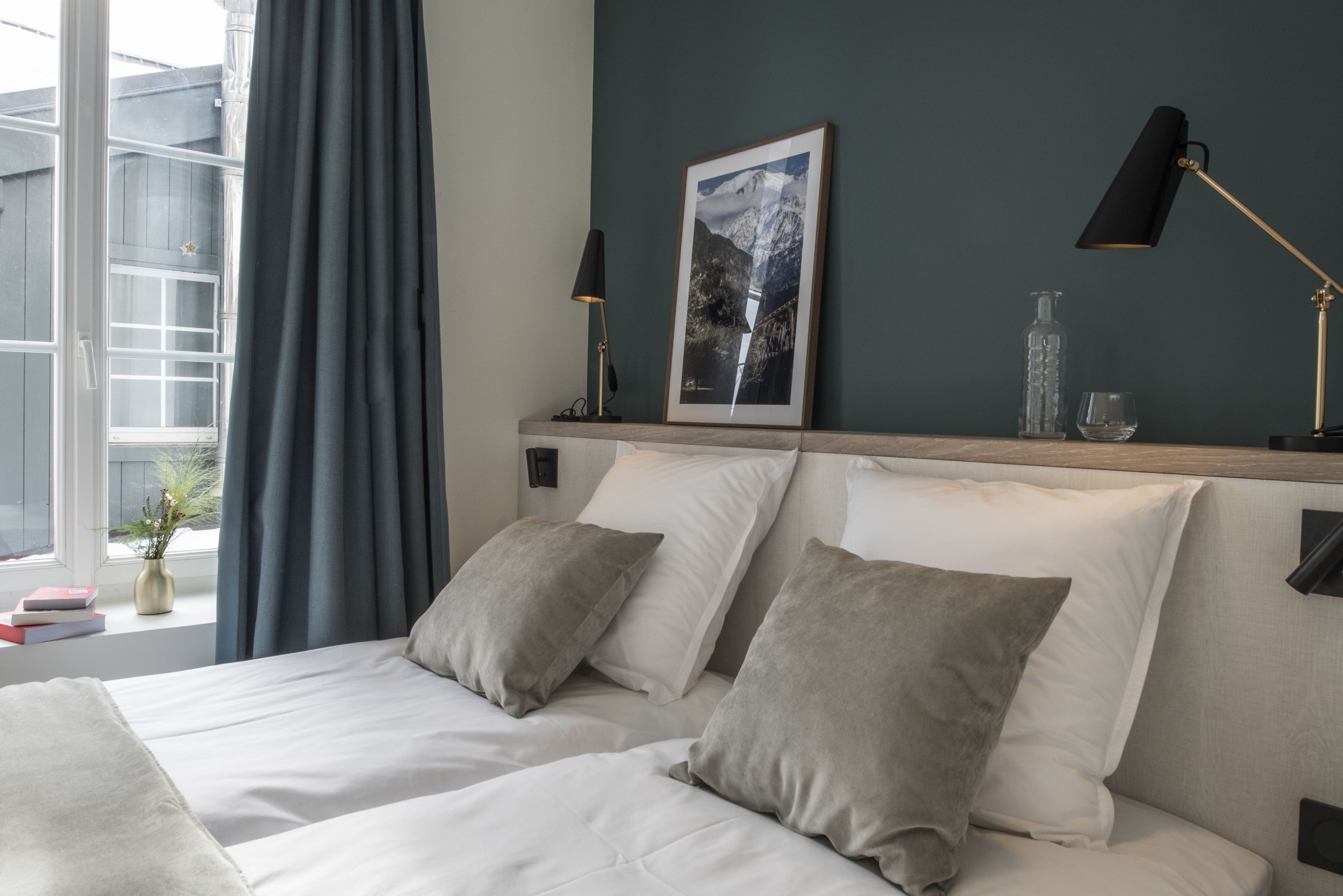 Una cama bien hecha con dos almohadas, un cuadro en la cabecera, una lámpara de noche y una ventana con cortinas.
