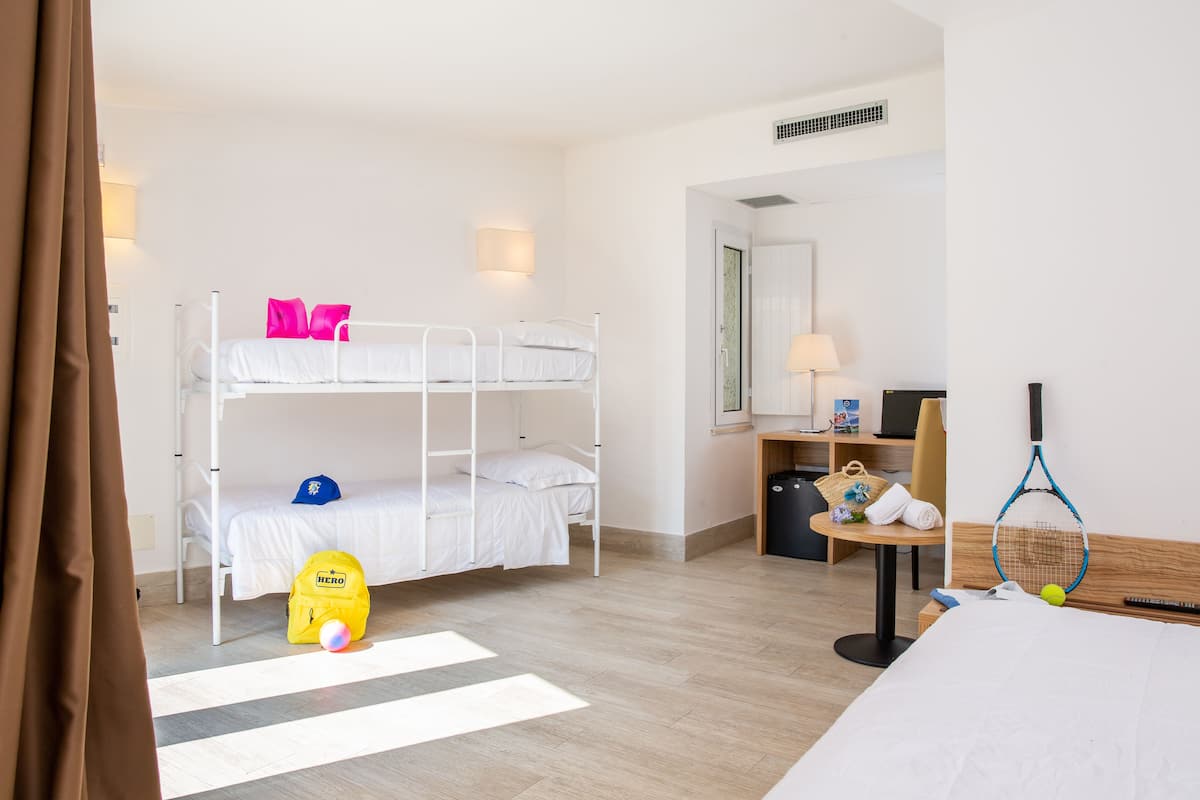 Une chambre d’hôtel avec des lits superposés, un bureau avec une lampe, une chaise, une petite table avec un sac et une raquette de tennis, et un lit avec une couverture.