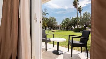 Un balcón abierto con sillas de mimbre y una mesa, con vista a un césped cuidado con palmeras y un edificio al fondo.