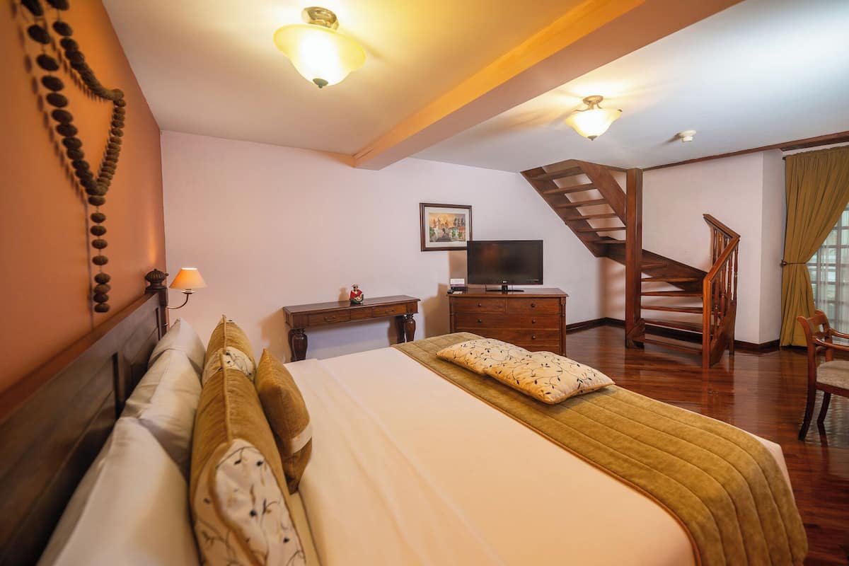 A hotel room with a bed, a television, a dresser, a desk, a chair, a staircase, and a wall-mounted picture.