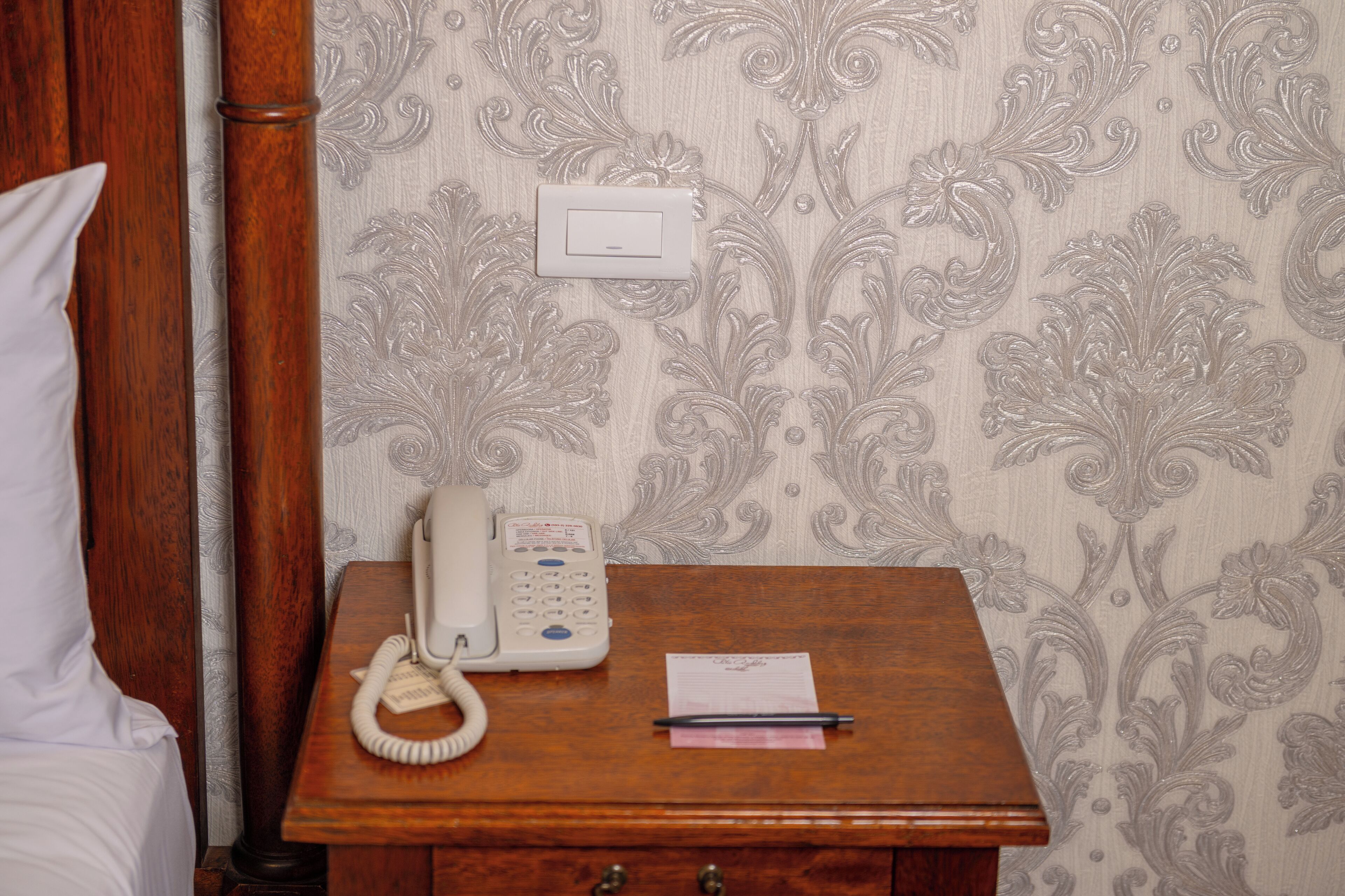 A wooden bedside table with a white telephone, a pen, and a piece of paper.