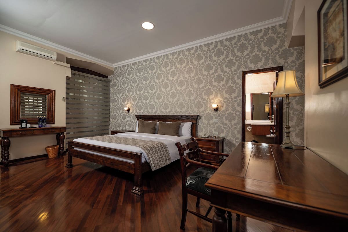 A hotel room with a wooden bed, a desk, a chair, a lamp, a mirror, and an air conditioning unit.