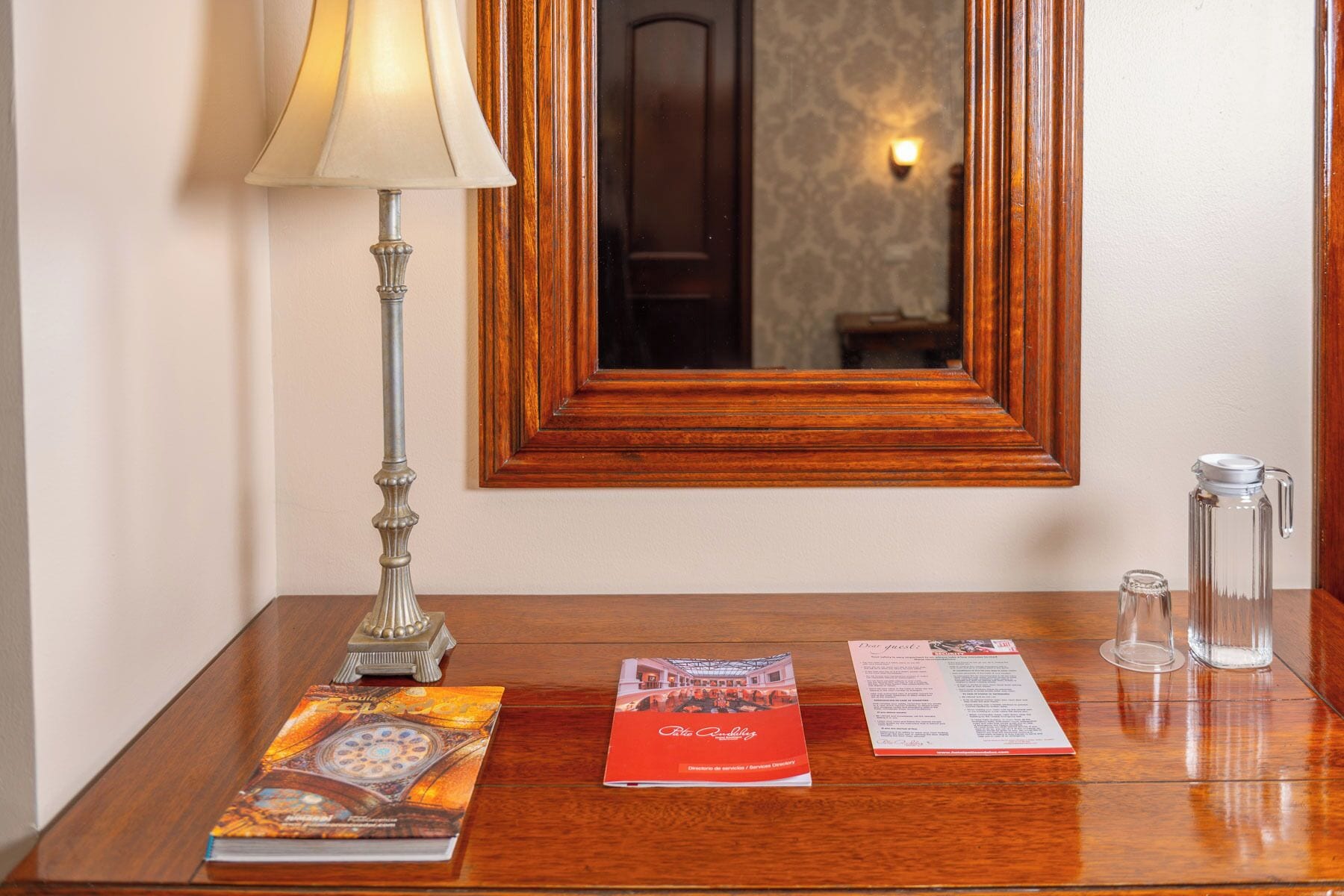 A wooden desk with a lamp, a mirror, a book, a brochure, and a glass pitcher.