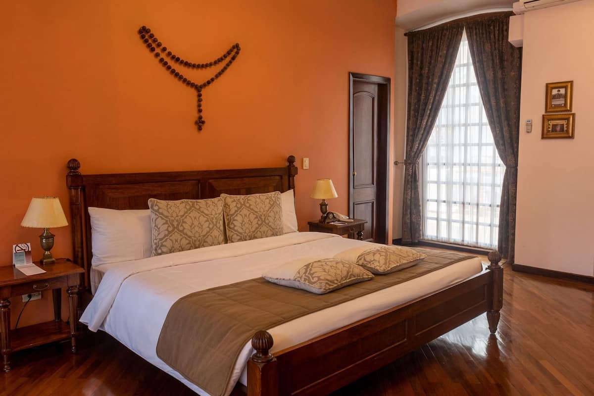 A neatly made bed with a wooden headboard, two bedside tables with lamps, a window with curtains, and a wall-mounted decorative item.