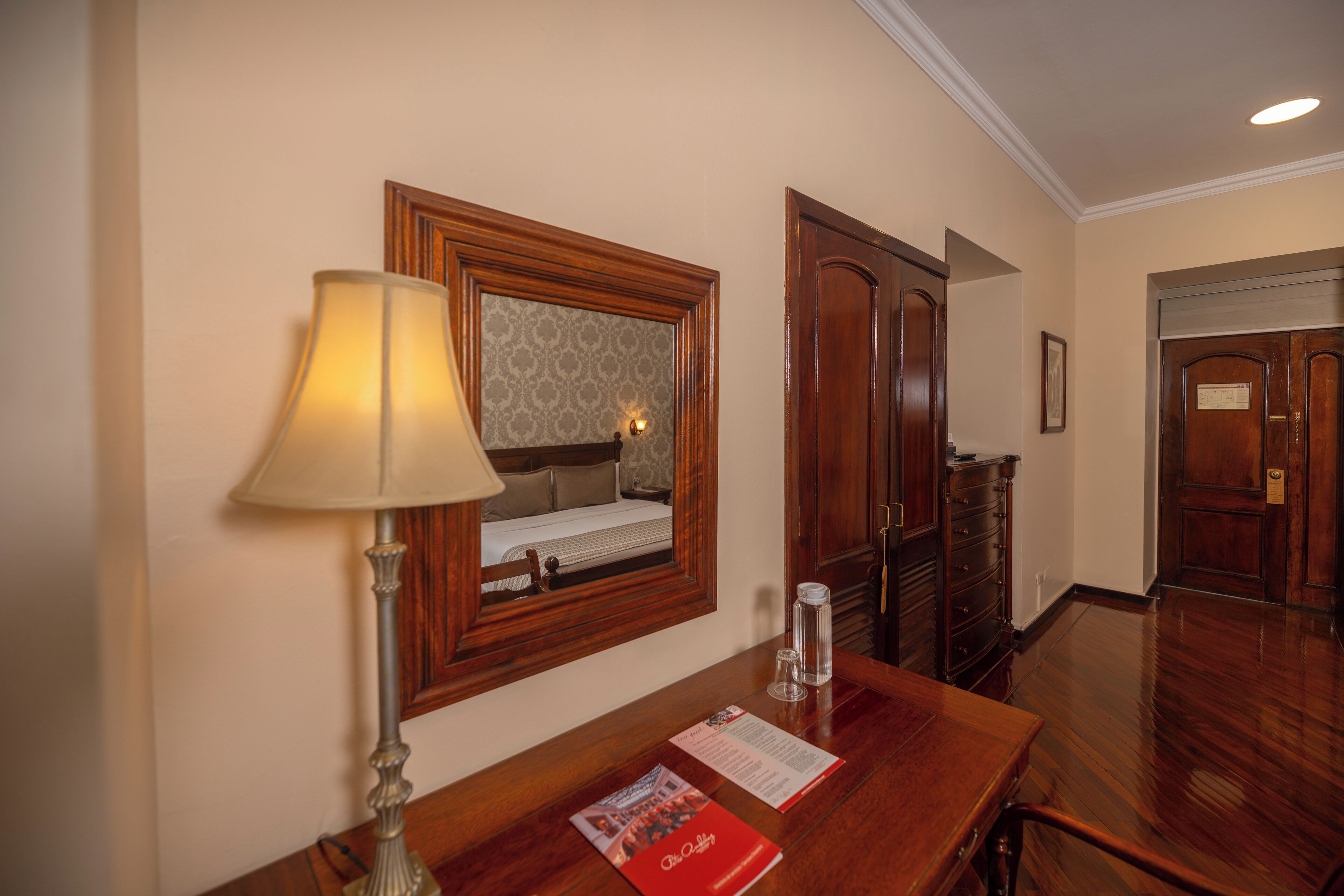 A hotel room with a wooden desk, a lamp, a framed mirror, a dresser, and a door.