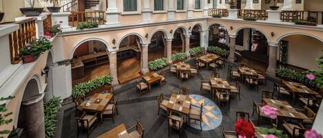 Un amplio patio interior con varias mesas y sillas, rodeado de arcos y columnas, con plantas en macetas y una escalera.