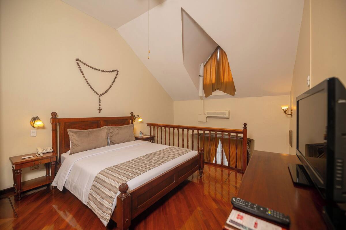 A bedroom with a wooden bed, a nightstand, a wall-mounted lamp, a television, and a ceiling light fixture.