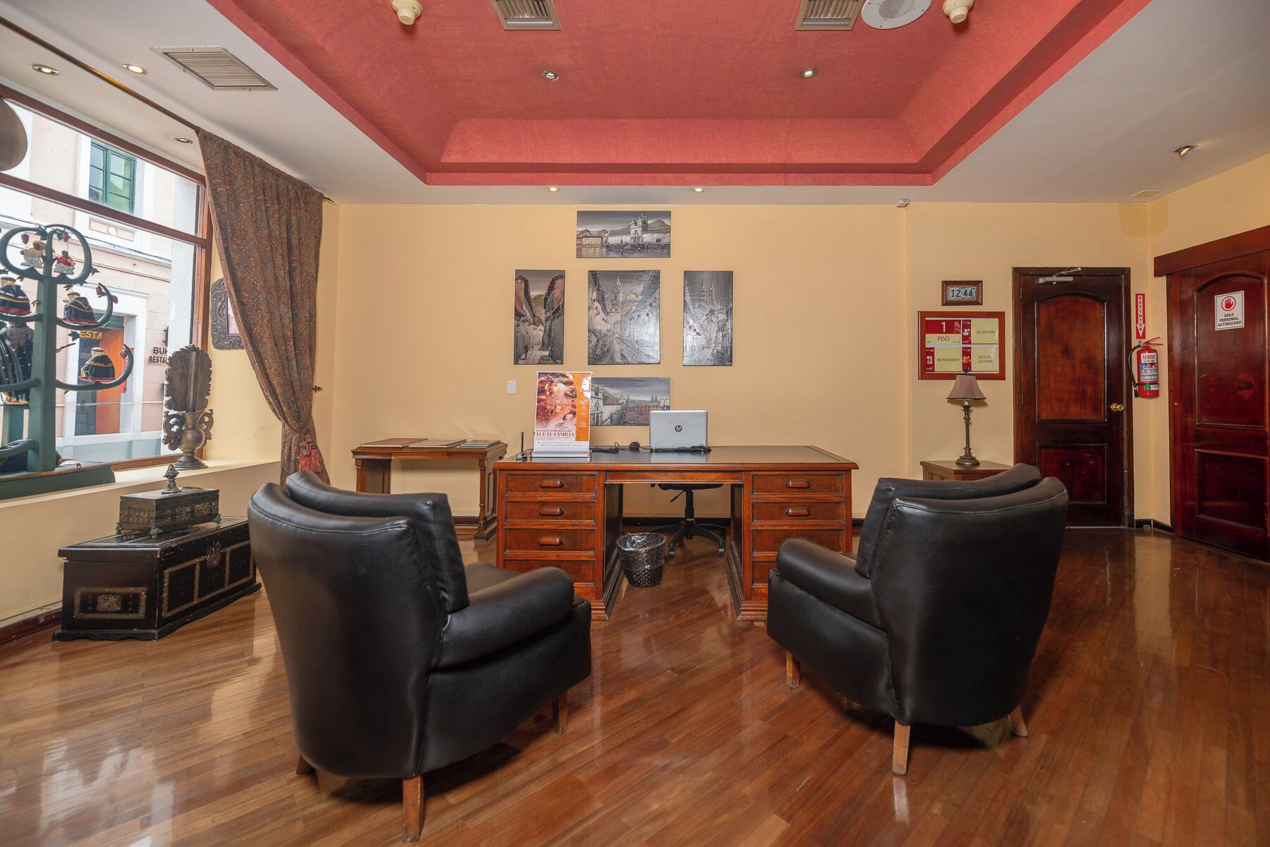 Habitación de hotel con escritorio de madera, dos sillones de cuero negro, una laptop y una ventana con vista a la calle.