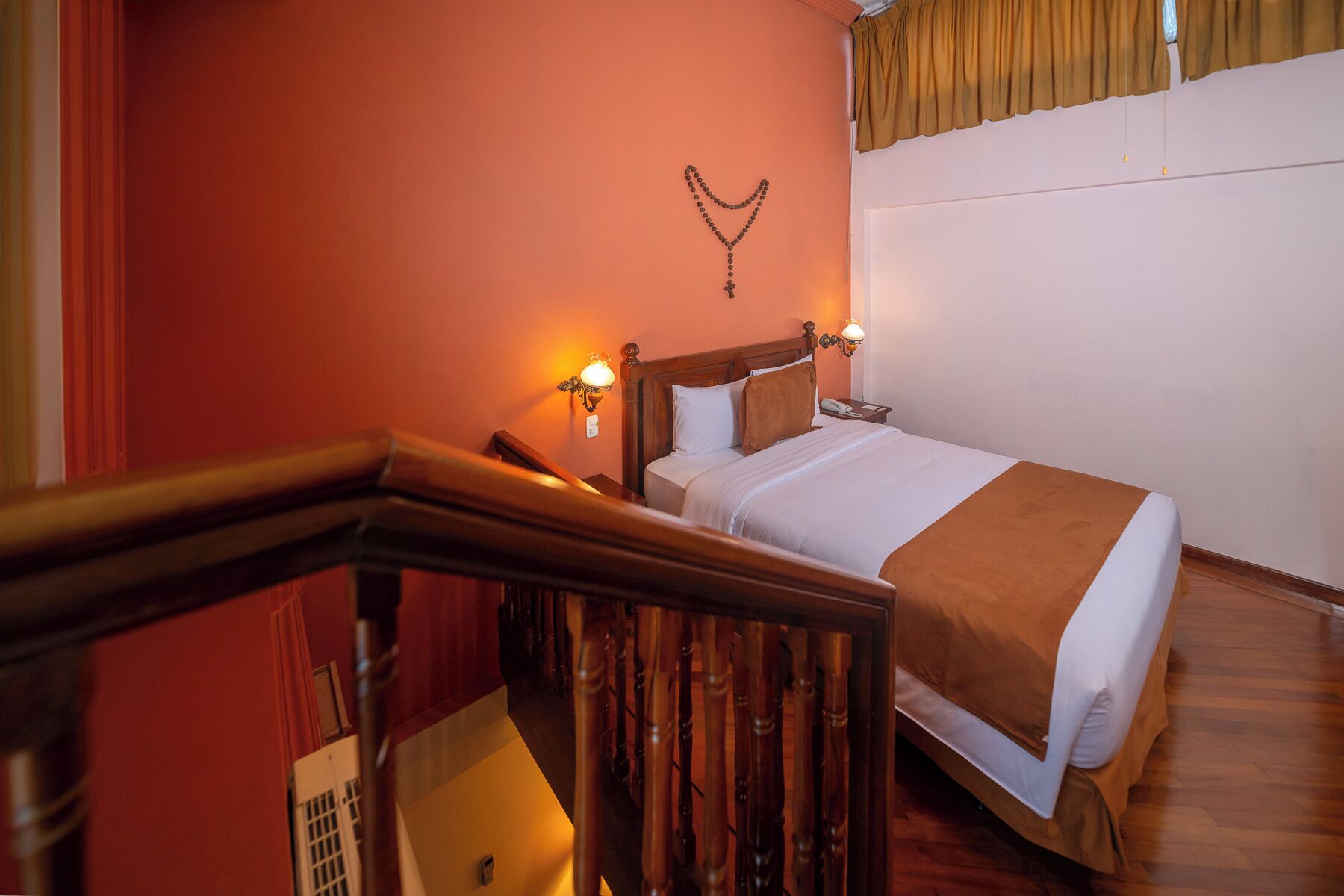 Una habitación de hotel con una cama de madera, dos lámparas de noche, un crucifijo en la pared y una ventana con cortinas.