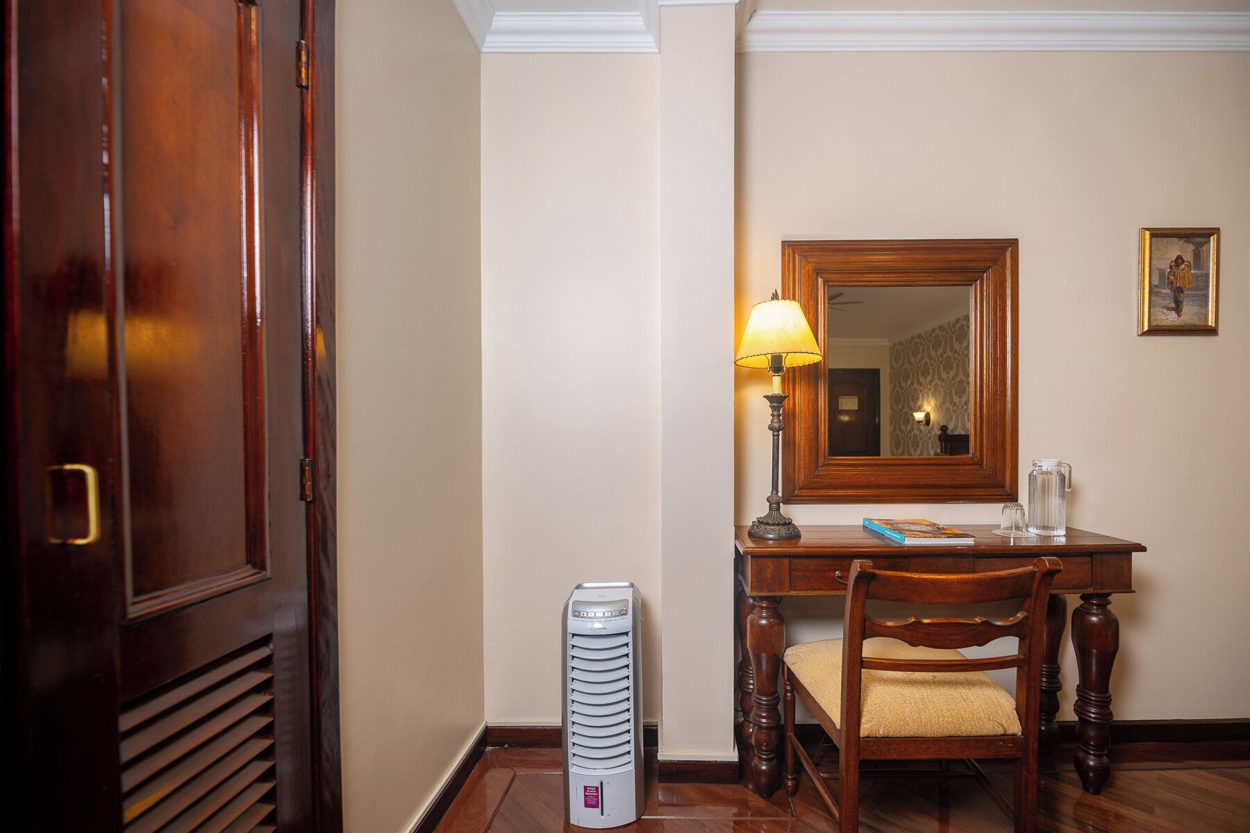 Habitación de hotel con un armario de madera, un escritorio con lámpara, una silla, un espejo y un calentador.