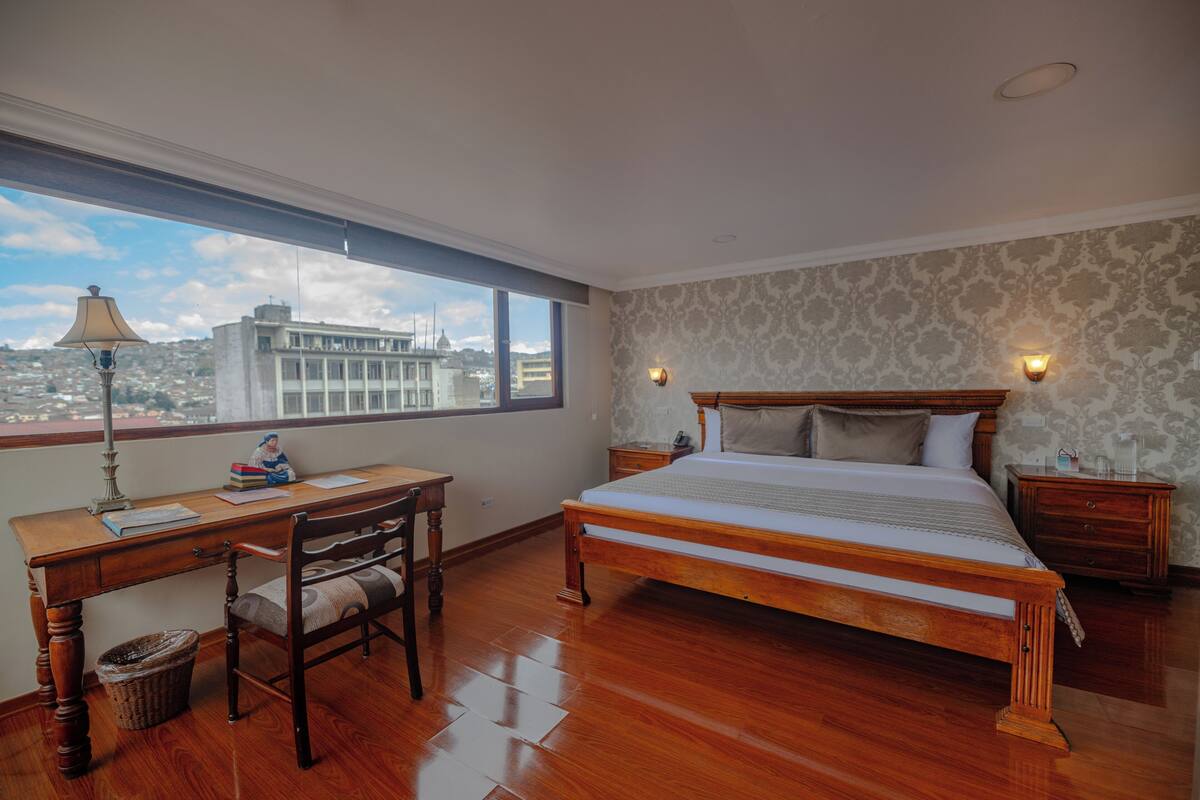 A bedroom with a wooden bed, a desk with a lamp, and a view of the city through a large window.