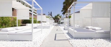 A pathway leading to white outdoor seating areas with cushions, surrounded by white pebbles, under a clear sky.