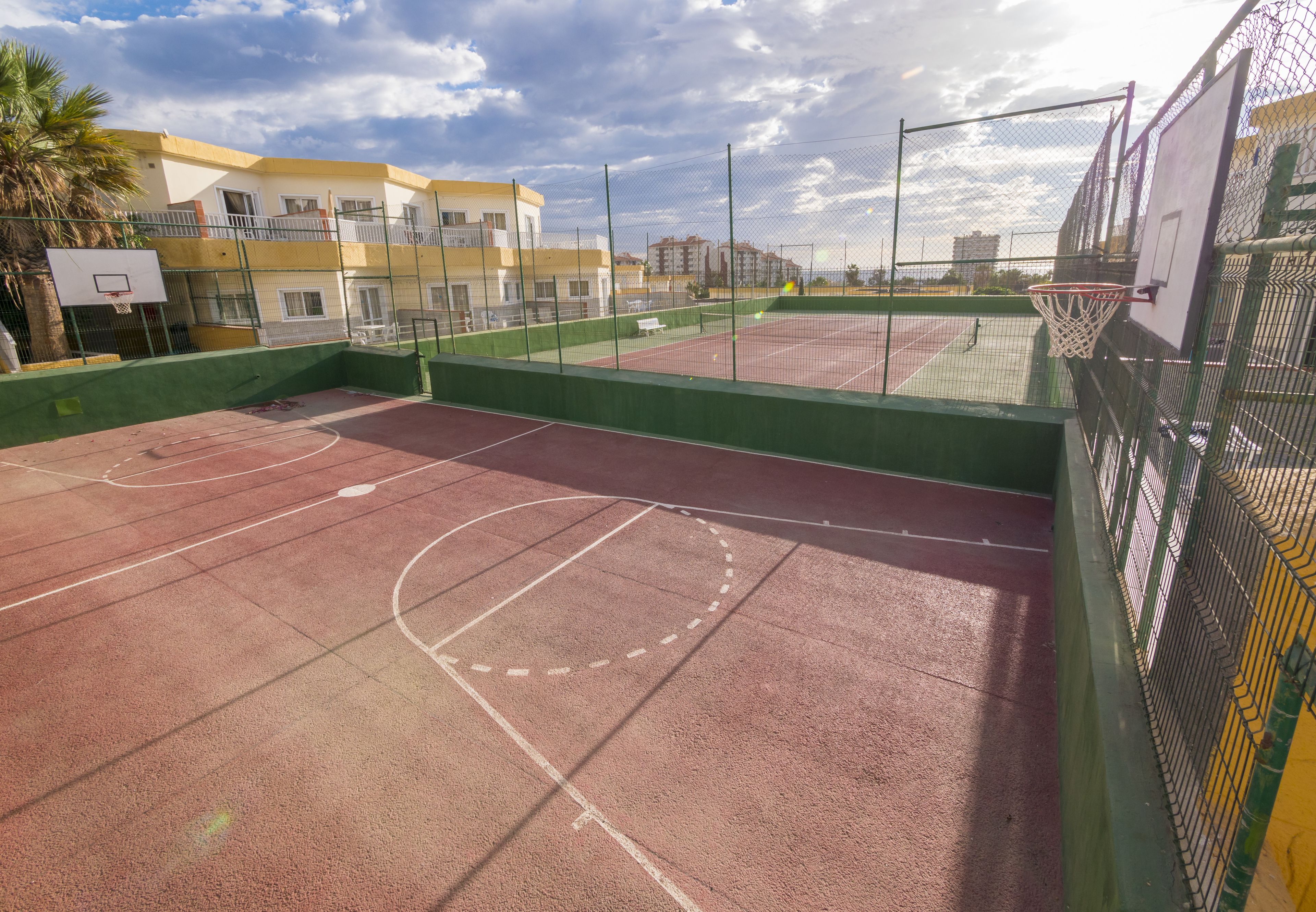 Una cancha de baloncesto al aire libre con una valla verde, un aro de baloncesto y una banca.