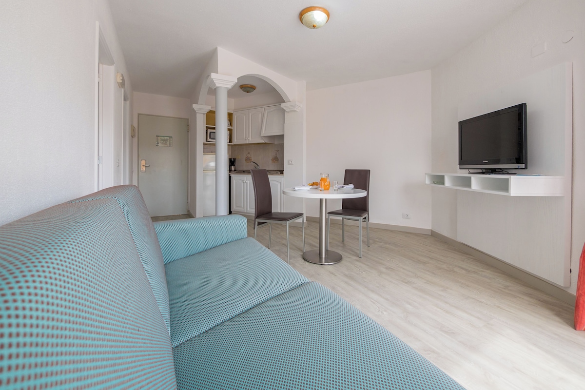 Una sala de estar moderna con un sofá, una mesa de comedor con sillas, una zona de cocina y un televisor montado en la pared.