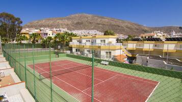 A tennis court with a green fence, surrounded by buildings with balconies and solar panels.