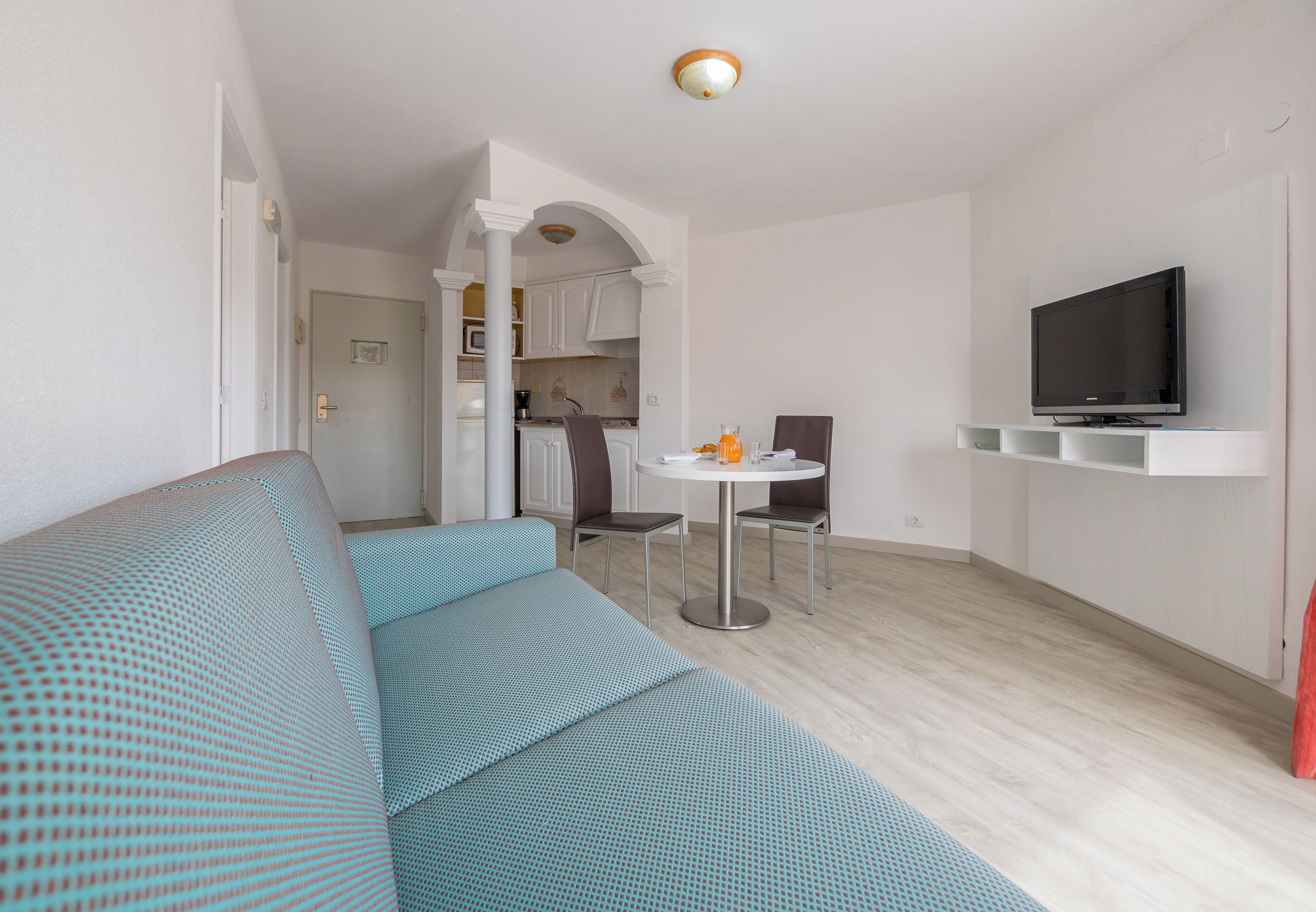 A modern living room with a sofa, a dining table with chairs, a kitchen area, and a mounted TV.