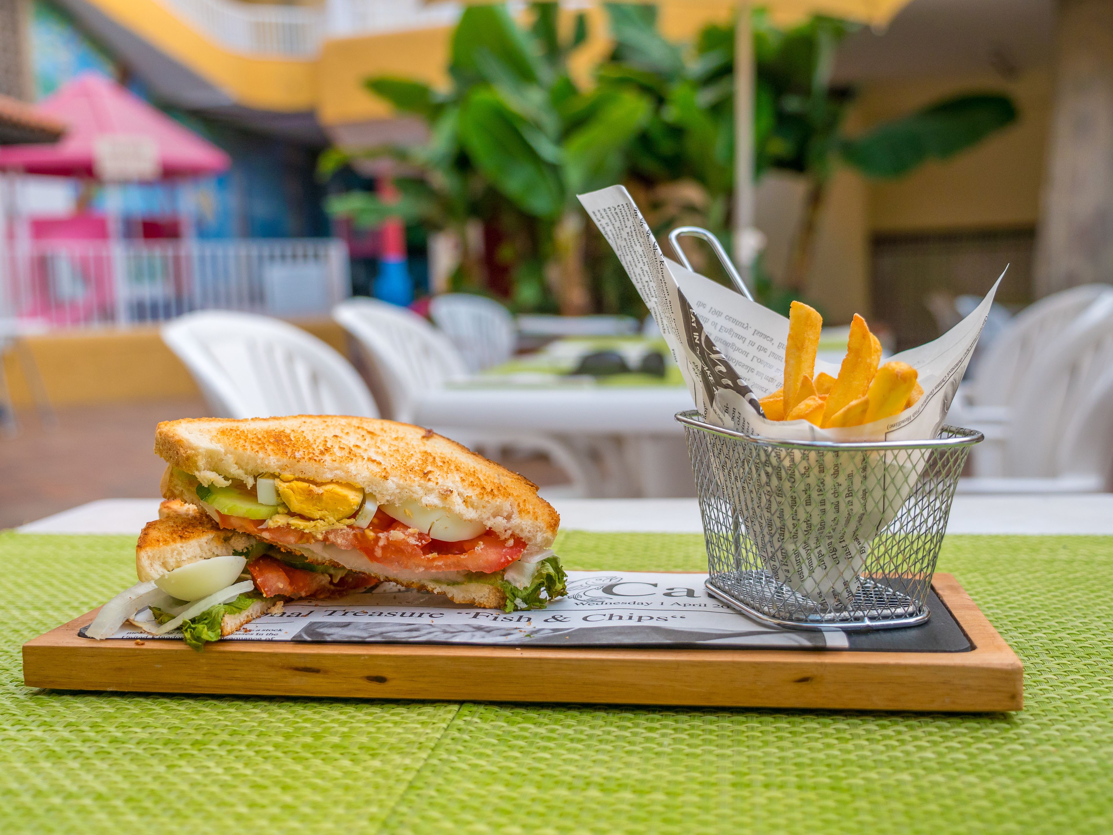 Un sándwich sobre una tabla de madera, con un periódico debajo y una cesta de papas fritas a un lado.