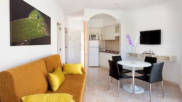 A modern living room with a brown sofa and yellow cushions, a dining area with a white table and black chairs, a kitchen with white cabinets, and a flat-screen TV mounted on the wall.