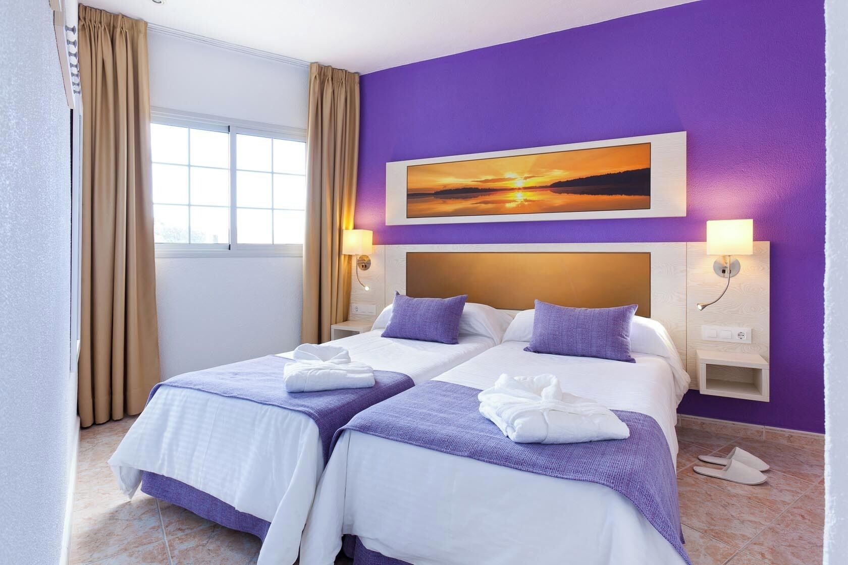 A hotel room with two beds, a large window, and a sunset painting on the wall.