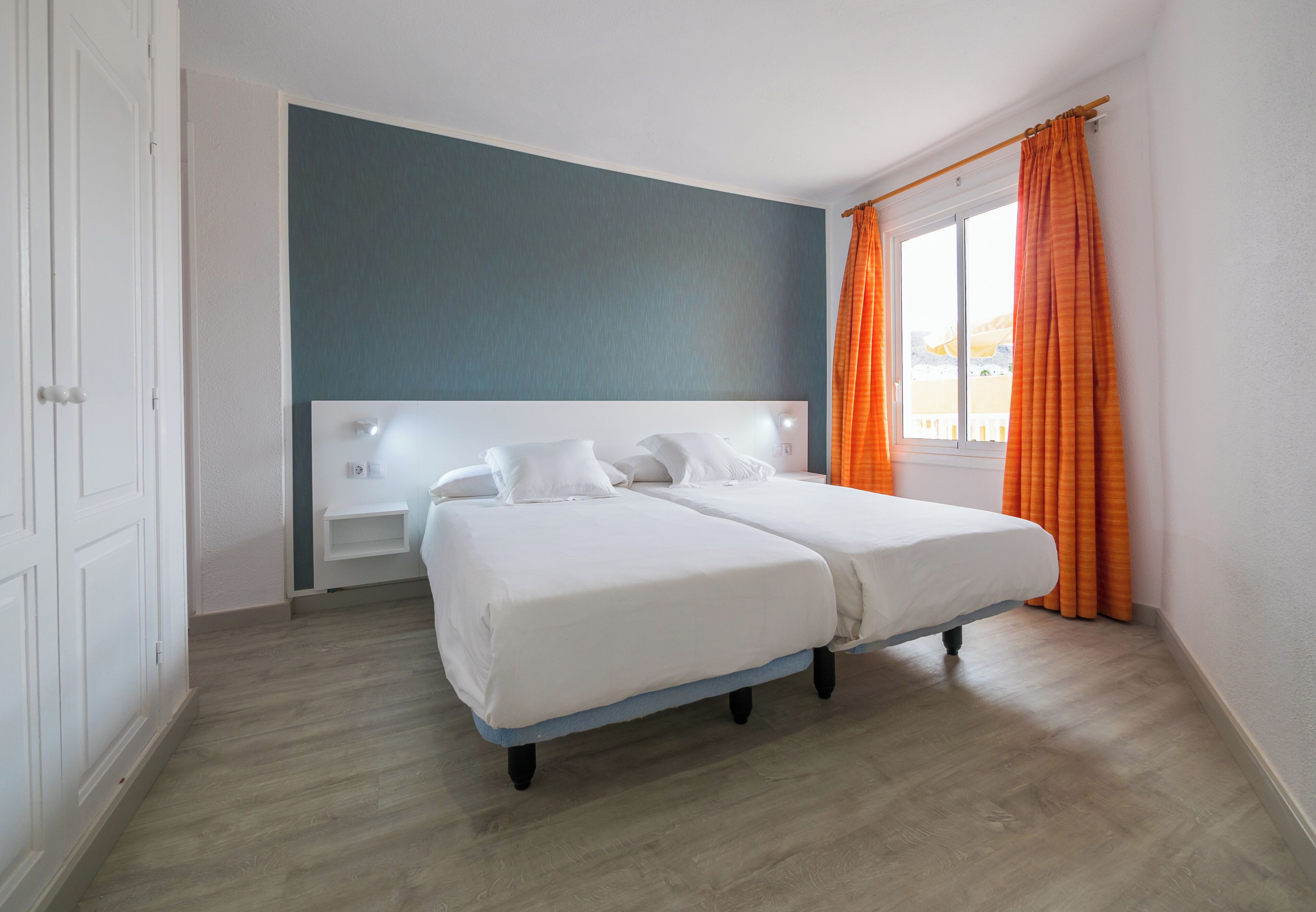 A double bed with white bedding, a bedside table, a blue accent wall, and an orange curtain.