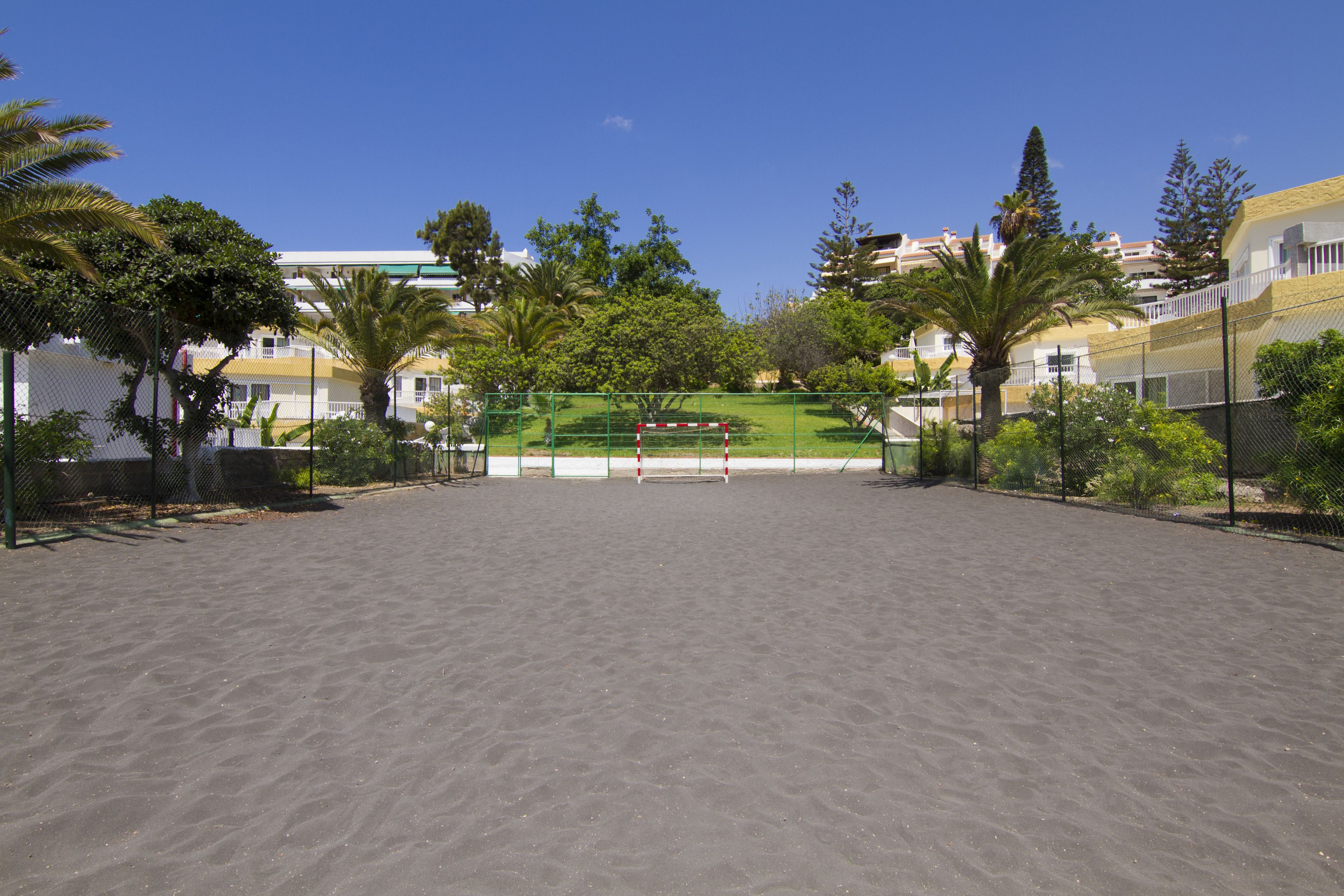 A sandy outdoor area with a volleyball net, surrounded by greenery and residential buildings.