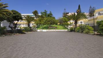 A sandy outdoor area with a volleyball net, surrounded by greenery and residential buildings.