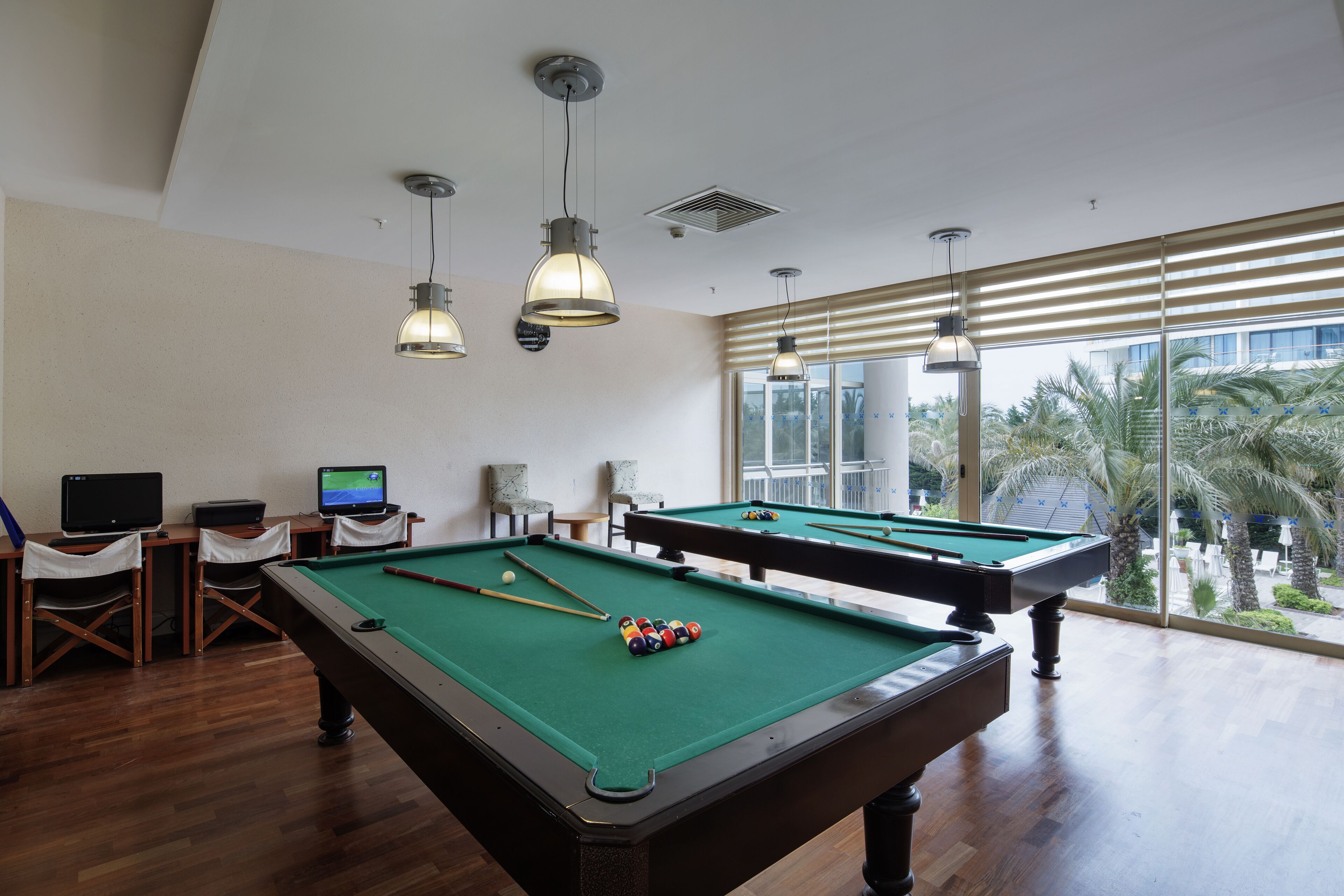 A pool table with a green felt surface, two pool cues, and a set of balls. There are two chairs and a small table with a laptop and a monitor. The room has wooden flooring and large windows with blinds.