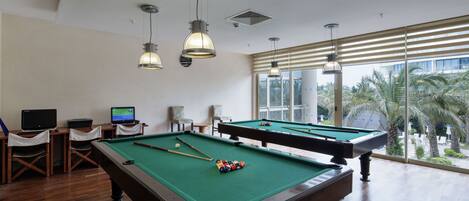 A pool table with a green felt surface, two pool cues, and a set of balls. There are two chairs and a small table with a laptop and a monitor. The room has wooden flooring and large windows with blinds.