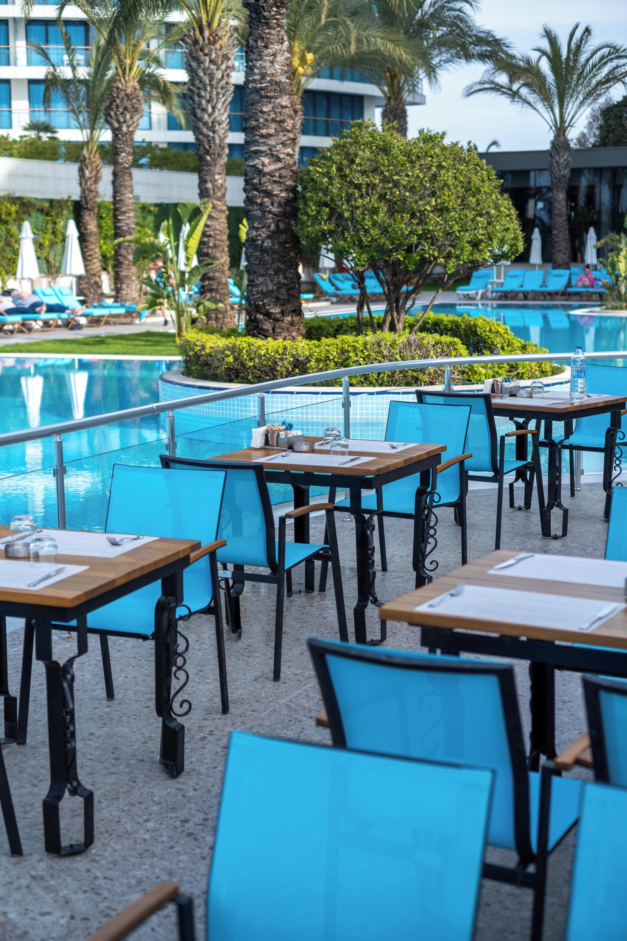 A poolside dining area with tables and chairs, a pool, and palm trees.